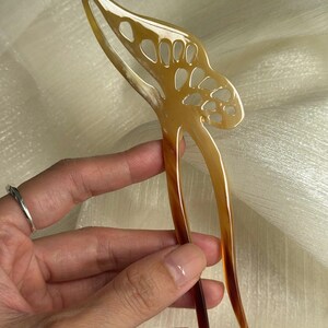 May include: A decorative hair pin with a butterfly design. The pin is made of a light brown material with a gradient of darker brown at the tips. The butterfly design has cut-out details.
