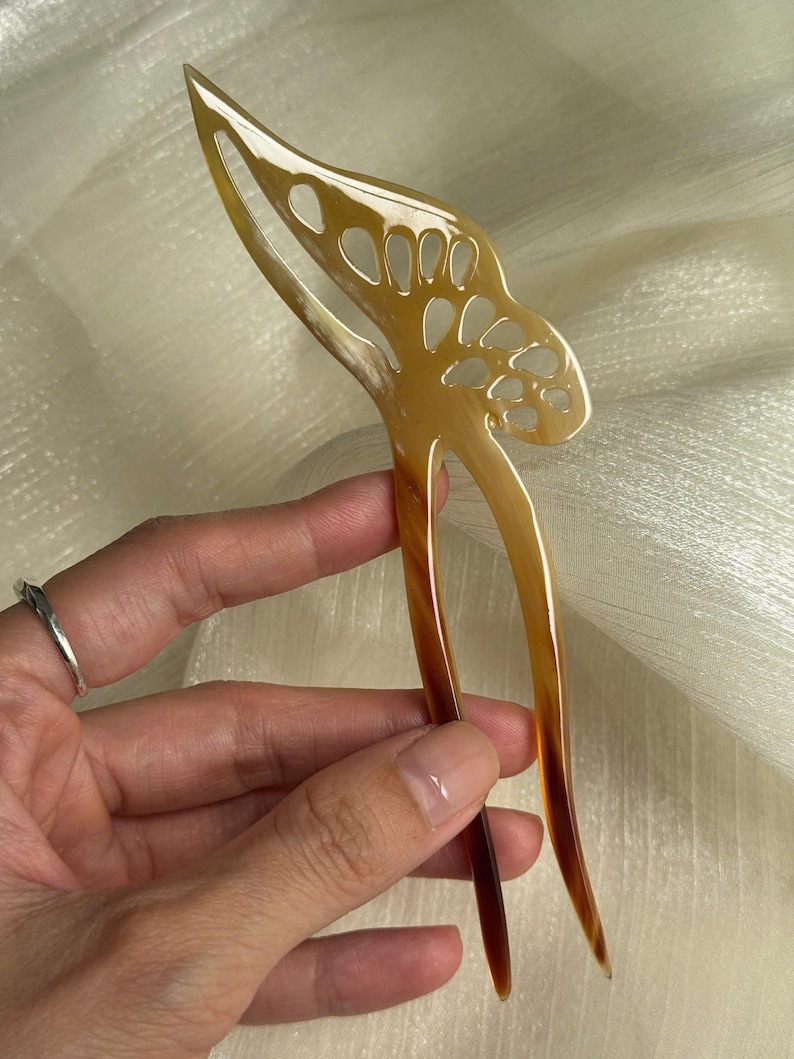 May include: A decorative hair pin with a butterfly design. The pin is made of a light brown material with a gradient of darker brown at the tips. The butterfly design has cut-out details.