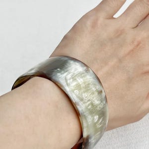 Pu&ograve; includere: Un bracciale rigido largo e liscio caratterizzato da un motivo traslucido e marmorizzato con tonalit&agrave; miste di crema, grigio chiaro e marrone scuro. Il bracciale presenta una finitura lucida e una struttura curva e solida.