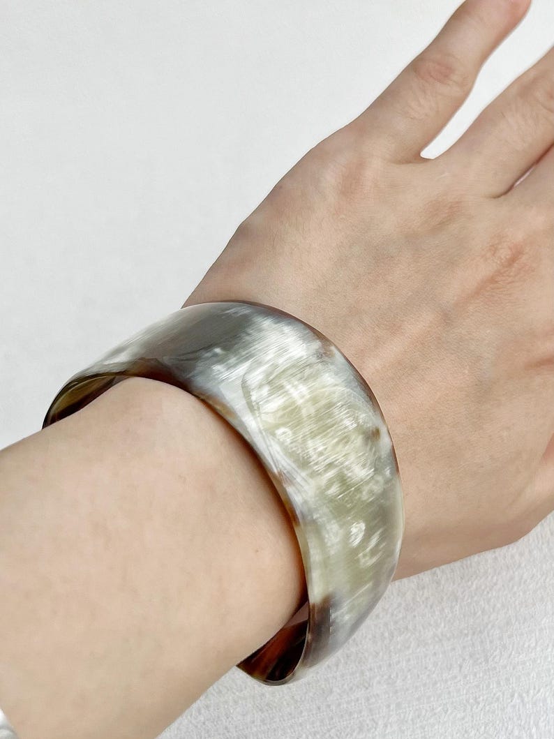 Pu&ograve; includere: Un bracciale rigido largo e liscio caratterizzato da un motivo traslucido e marmorizzato con tonalit&agrave; miste di crema, grigio chiaro e marrone scuro. Il bracciale presenta una finitura lucida e una struttura curva e solida.