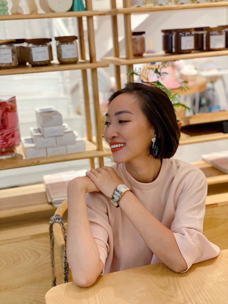 Pu&ograve; includere: Un top a maniche corte rosa chiaro indossato con un ampio bracciale a polsino con motivo marmorizzato nei toni del marrone, bianco e grigio, e orecchini pendenti con un design geometrico scuro e multicolore con una parte superiore in metallo argentato.