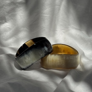 Pu&ograve; includere: Due bracciali rigidi larghi con una finitura liscia e lucida. Uno presenta un motivo marmorizzato nei toni del nero profondo e del bianco traslucido, mentre l'altro mostra una calda tavolozza di colori color miele e crema semitrasparenti.