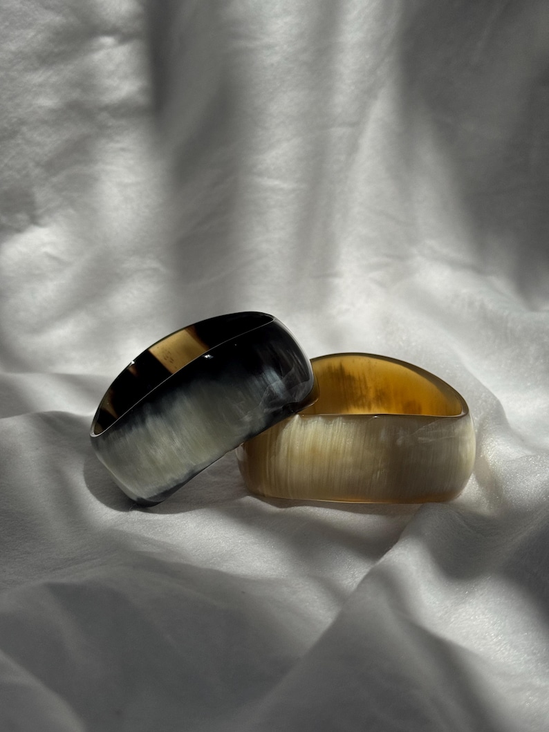 Pu&ograve; includere: Due bracciali rigidi larghi con una finitura liscia e lucida. Uno presenta un motivo marmorizzato nei toni del nero profondo e del bianco traslucido, mentre l'altro mostra una calda tavolozza di colori color miele e crema semitrasparenti.