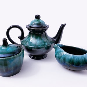 May include: A teal and black ceramic teapot, creamer and sugar bowl set. The teapot has a lid and a spout. The creamer and sugar bowl have lids.