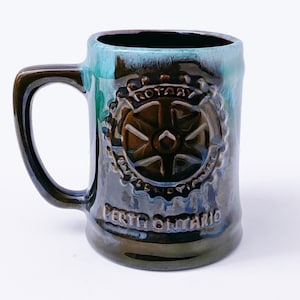 May include: A brown ceramic mug with a blue and green glaze. The mug features a rotary club logo with the text "Rotary" and "Perth Ontario".