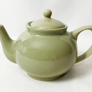 May include: A sage green ceramic teapot with a lid and a curved handle. The teapot has a rounded body, a spout, and a small knob on the lid. The teapot is designed for brewing tea.