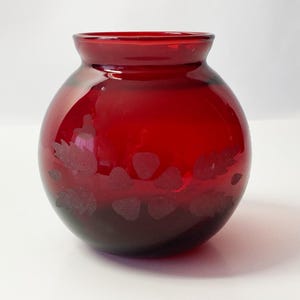 Peut inclure: Un petit vase rond en verre rouge avec un motif gravé de feuilles et de baies.