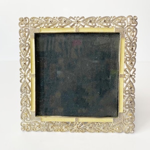May include: Square silver-toned picture frame with intricate filigree detailing. The frame has a pale yellow border around the dark interior. The decorative border features floral and heart-shaped elements, creating an elegant, vintage aesthetic.