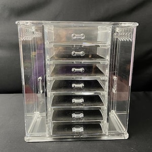 May include: Clear acrylic jewelry organiser with multiple drawers and side compartments. The organiser has a rectangular shape with a series of pull-out drawers of varying sizes. The side compartments have a hanger-like design. The clear material allows for easy viewing of contents.