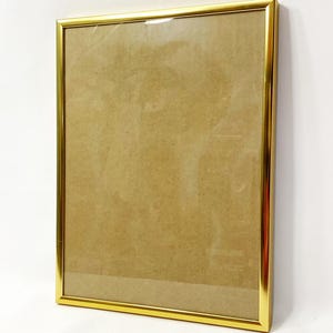 May include: A rectangular gold picture frame with a clear glass front. The frame has a shiny, metallic finish and is empty, with a brown backing visible through the glass. The frame is leaning against a white surface.