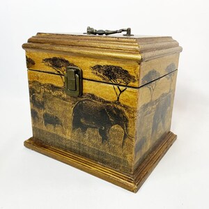 May include: A wooden box with a hinged lid, depicting a hand-painted African savanna scene with elephants and trees. The box has a brass clasp and handle, and a warm, golden-brown finish.
