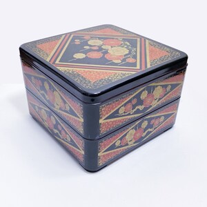 May include: A black, two-tiered Japanese bento box with a square shape. The box is decorated with floral and geometric patterns in red, gold, and black. The top lid features a diamond-shaped design with flowers.