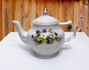 May include: A white ceramic teapot with a floral design featuring yellow and purple flowers. The teapot has a gold trim around the lid and spout.