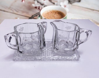 May include: A clear glass sugar bowl and creamer set with a decorative etched design. The set is on a matching clear glass tray.