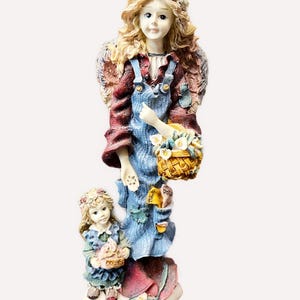 May include: A decorative angel figurine with a floral crown and wings, wearing a red jacket and blue dungarees. The angel holds a woven basket of flowers and stands with a smaller child figurine. The base is a cloud-like shape.