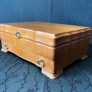 Peut inclure: Une boîte à bijoux en bois de couleur marron clair. La boîte est de forme rectangulaire avec une base incurvée et un bord supérieur décoratif. Elle comporte deux petits tiroirs avec des poignées en forme d'anneau de couleur dorée.
