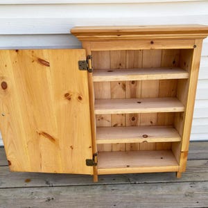 May include: A light-colored wooden cabinet with three shelves and a hinged door. The cabinet is made of natural wood with visible grain and knots. The door is open, revealing the interior shelves.