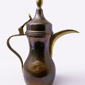 Könnte beinhalten: Eine arabische Kaffeekanne aus Messing mit einem Scharnierdeckel und einem Ausguss. Die Kanne hat ein dekoratives Design mit den Worten "Saudi-Arabien" an der Seite.