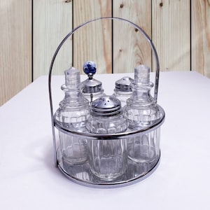 May include: A silver metal condiment holder with five glass jars. The jars have a clear glass body with a textured design. The holder has a round base and a handle.