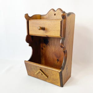 May include: A vintage wooden wall-mounted organiser with a drawer and a lower compartment. The organiser is made of stained wood with a decorative edge and a small wooden knob on the drawer. The lower compartment has a carved design.