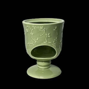 May include: A green ceramic candle holder with a floral design. The holder has a pedestal base and a cutout in the centre for a candle.