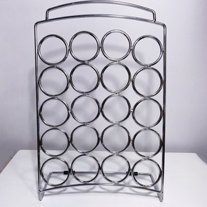 Peut inclure: Un porte-dosettes à café en métal chromé avec 20 fentes circulaires pour ranger les dosettes de café. Le porte-dosettes a une base incurvée et une poignée en haut.