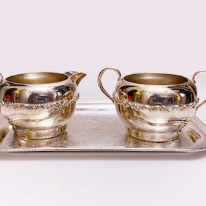 May include: A silver creamer and sugar bowl set on a silver tray. The creamer has a single handle and the sugar bowl has two handles. Both pieces have a decorative design around the rim.
