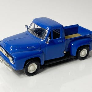 May include: A die-cast model of a classic blue pickup truck. The truck features a detailed front grille, side mirrors, and white-walled tires. The truck bed is a lighter shade of blue. A vintage collectible.