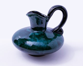May include: A small, green ceramic pitcher with a black handle. The pitcher has a glossy finish and a rounded body.