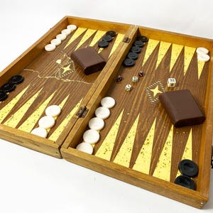 May include: A wooden backgammon board with a brown finish and gold and yellow accents. The board is open, revealing black and white game pieces, dice, and two brown cases. The game is set up and ready to play.