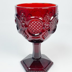 May include: A vintage ruby red glass goblet with a detailed pattern. The goblet has a textured design on the bowl and a geometric base. The stem is faceted, and the rim is smooth. The glass is a deep, rich red colour.