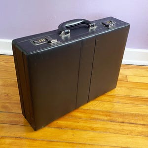 May include: A black leather briefcase with a handle and combination locks. The briefcase is rectangular and sits on a wooden floor. The handle and locks are silver-toned.