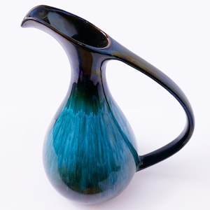May include: A ceramic jug with a long, curved handle and a spout. The jug has a teardrop shape and is glazed in shades of teal, blue, and black. The handle and spout are black. The jug is on a white background.