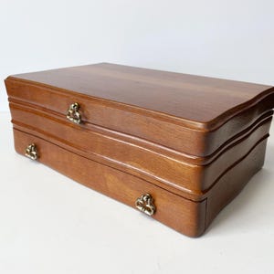 Peut inclure: Une boîte à bijoux en bois avec une finition marron chaud. La boîte a une forme rectangulaire avec un design incurvé sur le devant. Elle comporte deux tiroirs avec des poignées en métal ornées. La boîte est posée sur un fond blanc.