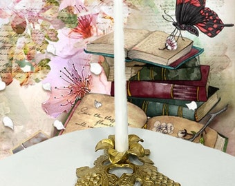 May include: A white candle sits in a gold metal candlestick with a grapevine design. The candlestick is on a white surface.