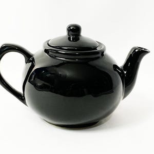 May include: A glossy black ceramic teapot with a rounded body, curved handle, and a spout. The lid has a small knob on top. The teapot is designed for brewing tea and has a classic, elegant appearance.