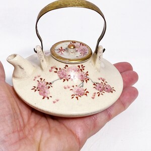 May include: A small white ceramic teapot with a gold handle and lid. The teapot is decorated with pink and red floral designs.