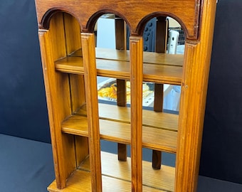 May include: A wooden wall shelf with three shelves and arched openings. The shelf is made of light brown wood and has a rectangular shape. The back of the shelf appears to be mirrored, reflecting the surroundings.