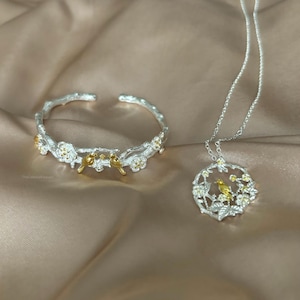 May include: A silver bracelet and necklace set featuring a delicate design of branches, flowers, and birds. The birds are accented with gold.