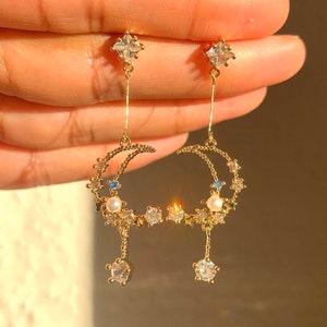 Puede incluir: Pendientes de oro en forma de media luna con estrellas colgantes y un acento de perla. Los pendientes están adornados con cristales transparentes y azules.