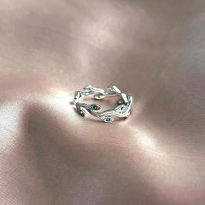 Op de afbeelding: Een zilveren ring met een delicaat takontwerp en kleine groene edelstenen. De ring ligt op een roze stoffen achtergrond.