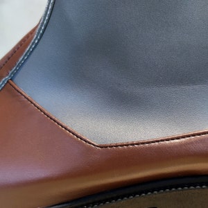 May include: Close-up of a brown leather boot with a black leather panel on the top. The boot has a stitched seam around the edge of the panel.