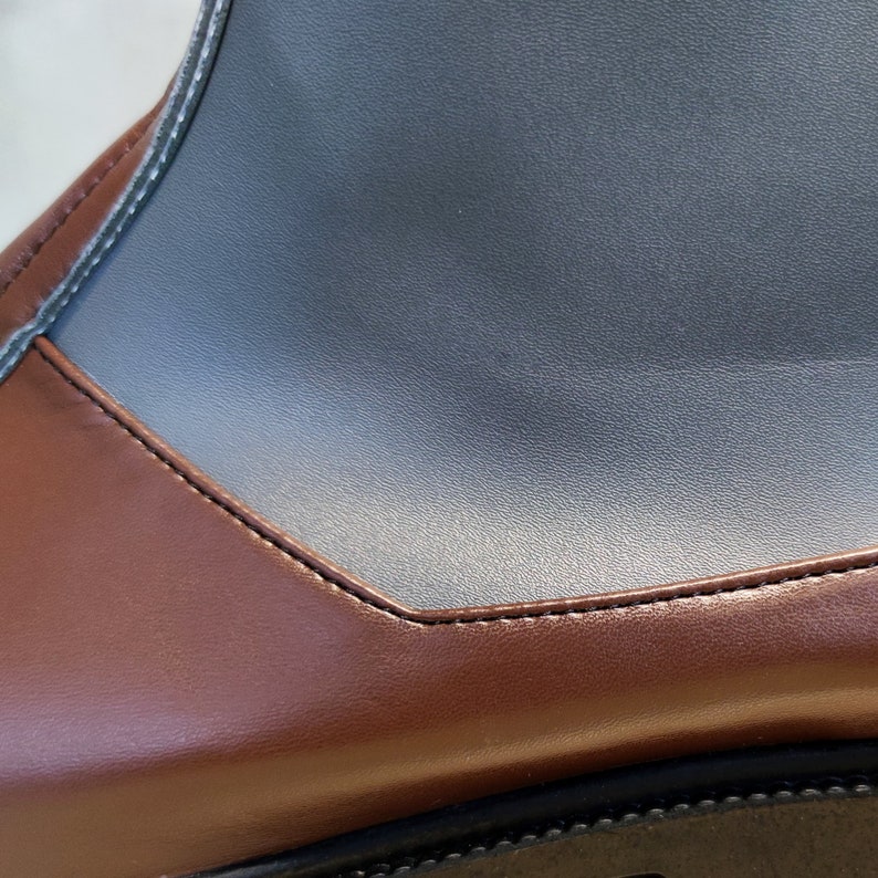May include: Close-up of a brown leather boot with a black leather panel on the top. The boot has a stitched seam around the edge of the panel.