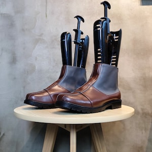 May include: A pair of brown leather boots with gray fabric panels and black boot shapers. The boots are sitting on a light wood table.