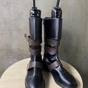 K&ouml;nnte beinhalten: Ein Paar schwarze Lederstiefel mit braunen Riemen und Schnallen. Die Stiefel haben eine spitze Zehenpartie und einen niedrigen Absatz. Sie werden von Schuhspannern gehalten.