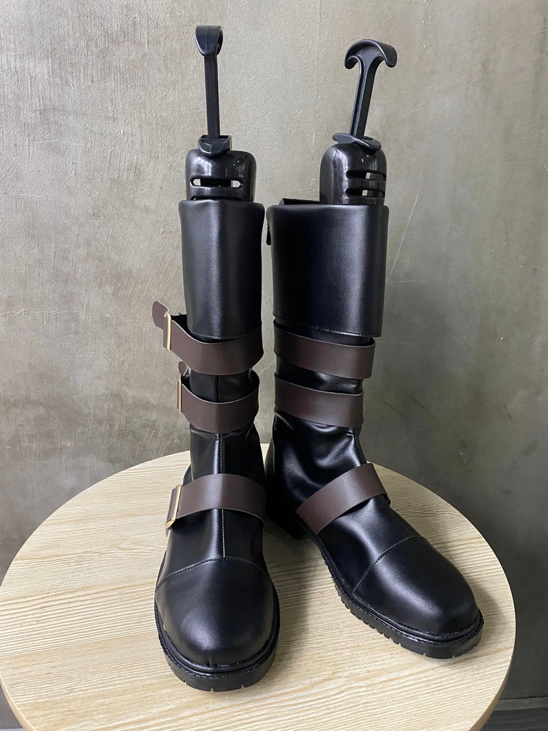 K&ouml;nnte beinhalten: Ein Paar schwarze Lederstiefel mit braunen Riemen und Schnallen. Die Stiefel haben eine spitze Zehenpartie und einen niedrigen Absatz. Sie werden von Schuhspannern gehalten.