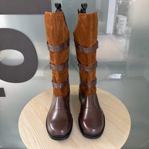 May include: A pair of knee-high brown leather boots with suede uppers. The boots have three brown leather straps with buckles and a black sole. The boots are displayed on a light brown wooden surface.