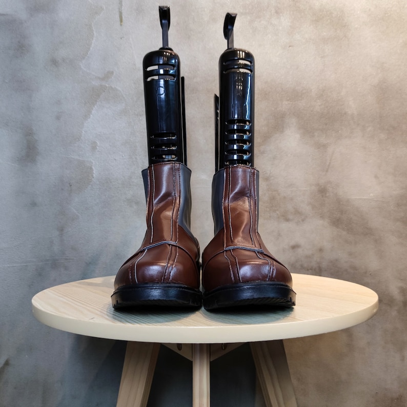 May include: A pair of brown leather boots with black shoe trees inside. The boots are sitting on a light wood table.