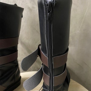 K&ouml;nnte beinhalten: Schwarze Lederstiefel mit braunen Riemen und einem Rei&szlig;verschluss auf der R&uuml;ckseite. Die Stiefel haben eine Schnalle an der Seite.
