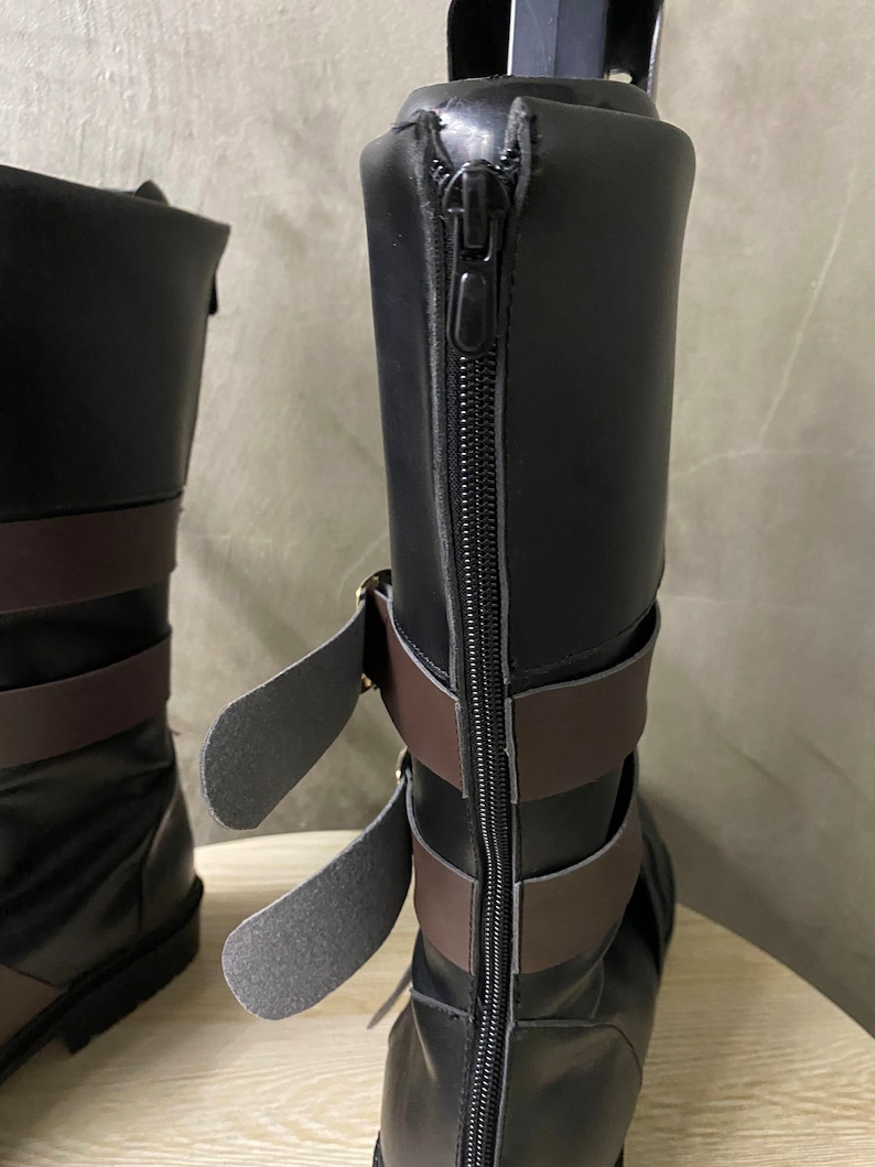 K&ouml;nnte beinhalten: Schwarze Lederstiefel mit braunen Riemen und einem Rei&szlig;verschluss auf der R&uuml;ckseite. Die Stiefel haben eine Schnalle an der Seite.